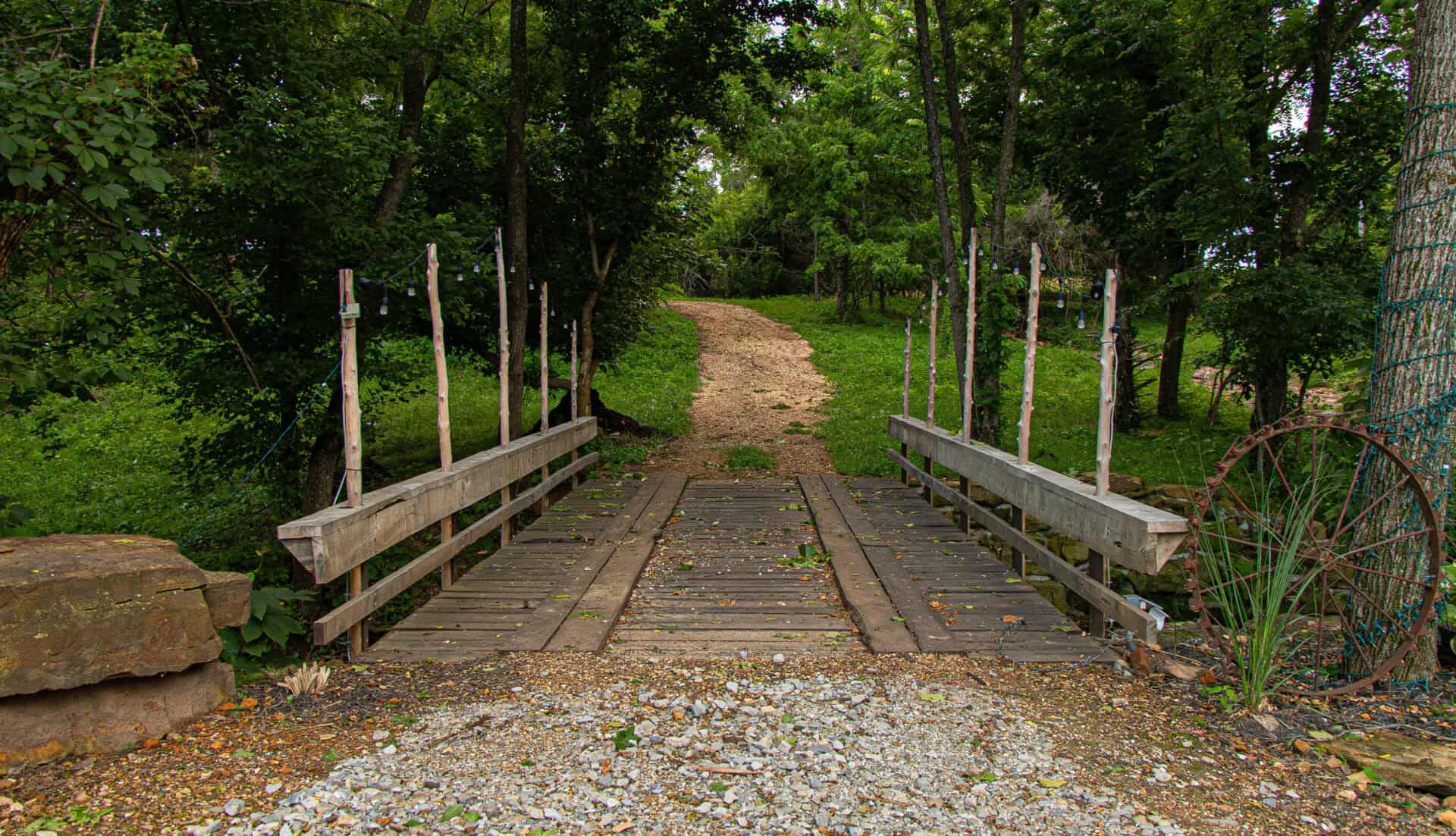 Uneven wooden footbridge over a small creek surrounded by lush green trees and foliage, leading to a rustic outdoor wedding or event venue in nature.