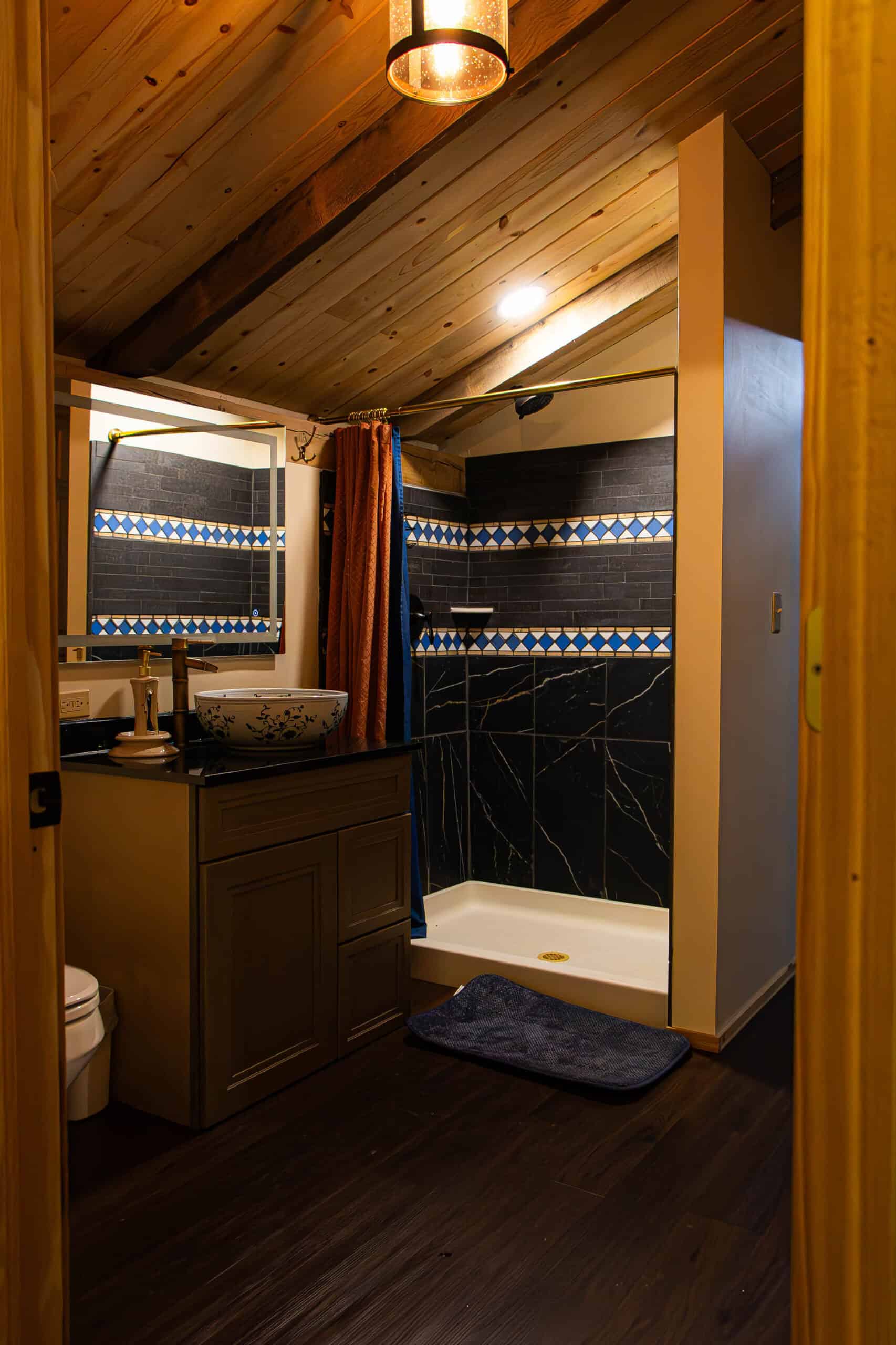 Cozy rustic bathroom at Belle Stone Venue featuring wooden ceiling, stylish black and blue tile accents, a modern vanity with a vessel sink, and a walk-in shower with dark marble tiles. Perfect for wedding and event accommodations.
