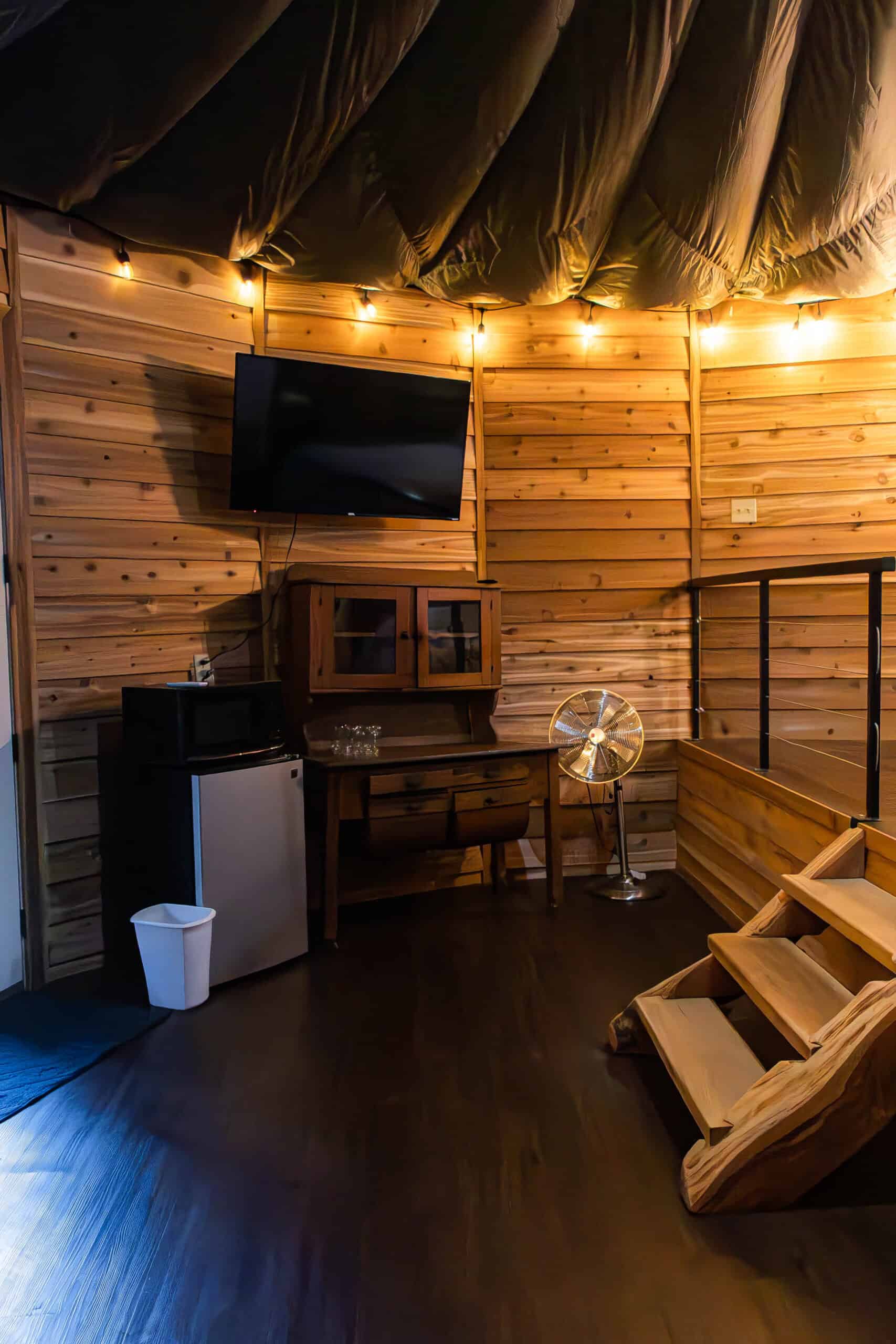 Cozy wooden interior with a mounted flat-screen TV, warm string lights, and rustic furniture at Belle Stone Venue, ideal for intimate gatherings or events with charming ambiance.