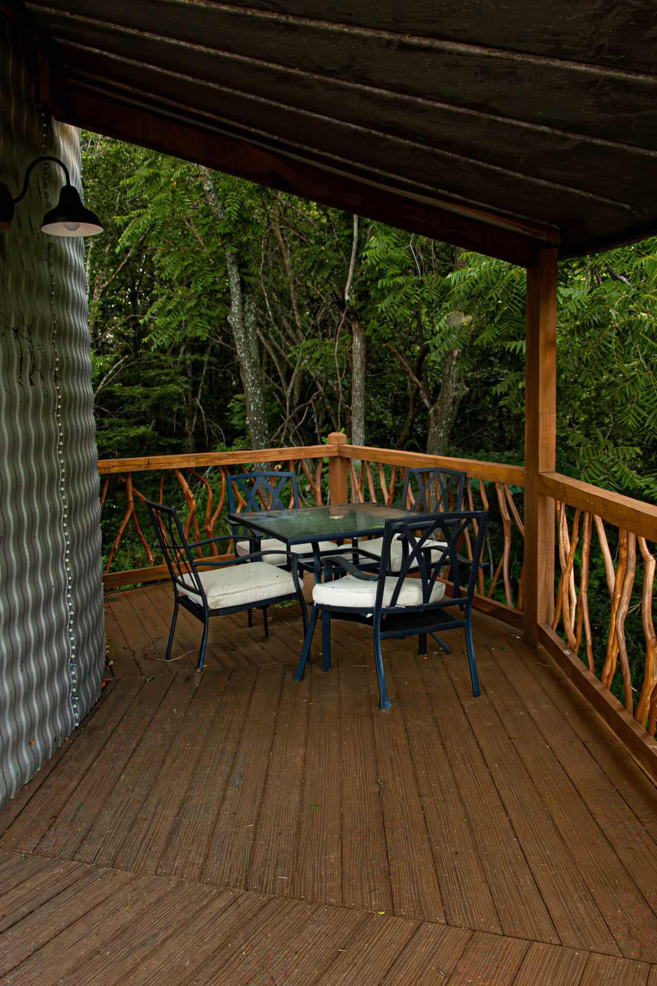 Rustic outdoor balcony with wooden deck, black metal chairs with white cushions, glass-top table, surrounded by lush green trees, ideal for outdoor wedding receptions and gatherings at Belle Stone Venue.