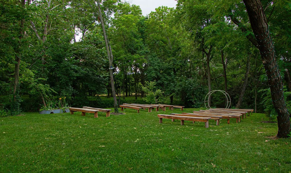 A peaceful outdoor wedding ceremony setup with wooden benches on lush green grass surrounded by trees at Belle Stone Venue.