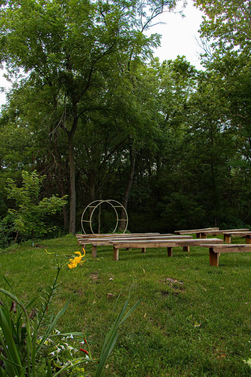 Serene outdoor wedding ceremony space at Belle Stone Venue with wooden benches, lush greenery, and a modern circular sculpture, perfect for garden-themed events or rustic weddings.