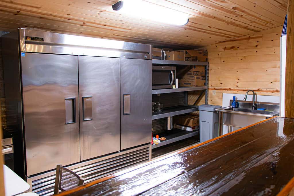 Stainless steel commercial refrigerator in a rustic event venue kitchen with wooden walls and ceiling.