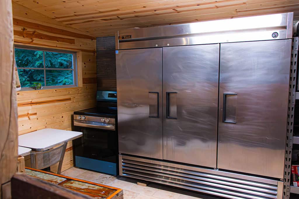 Modern commercial kitchen with large stainless steel refrigerator, black stove, wood-paneled walls, small window, and simple dining area at Belle Stone Venue.
