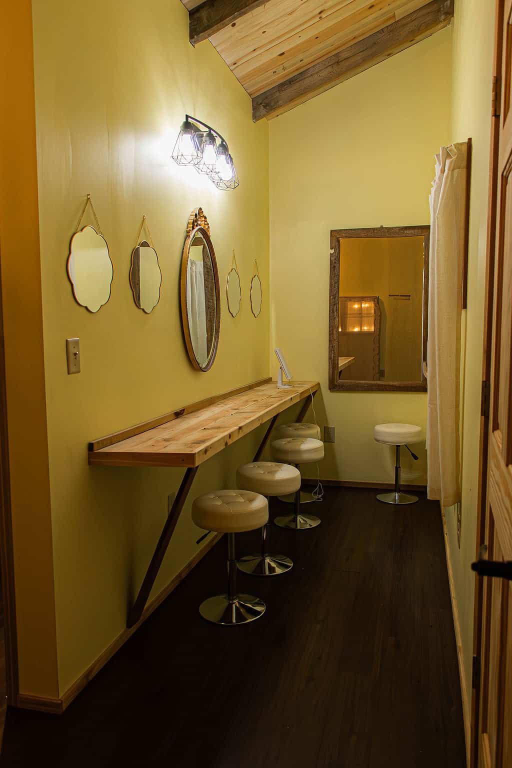 Cozy interior of Belle Stone Venue featuring a rustic wooden bar-height table with four upholstered bar stools, vintage mirrors on a warm yellow wall, and modern lighting, ideal for elegant events and gatherings.