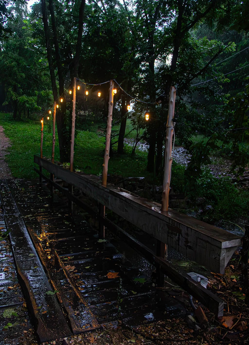String lights hanging over a wooden bridge in a lush, green outdoor venue at dusk, creating a cozy and enchanting atmosphere for events or gatherings.